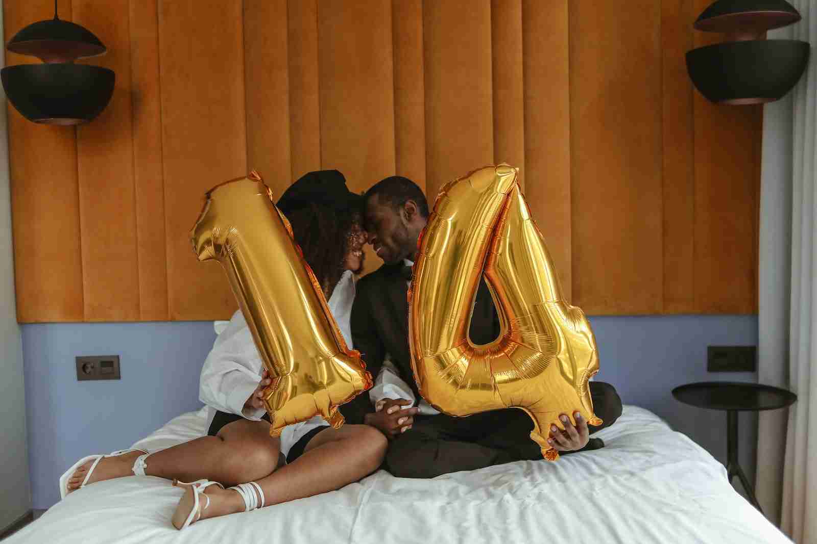 A Couple Sitting on the Bed While Holding 14 Number Balloon