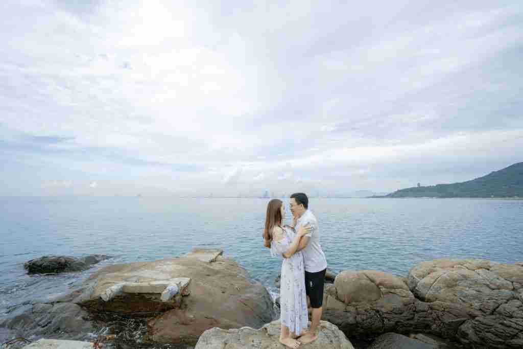 Side view full body of barefoot couple in summer clothes cuddling on big boulders on beach near sea in summer day under cloudy sky while looking at each other