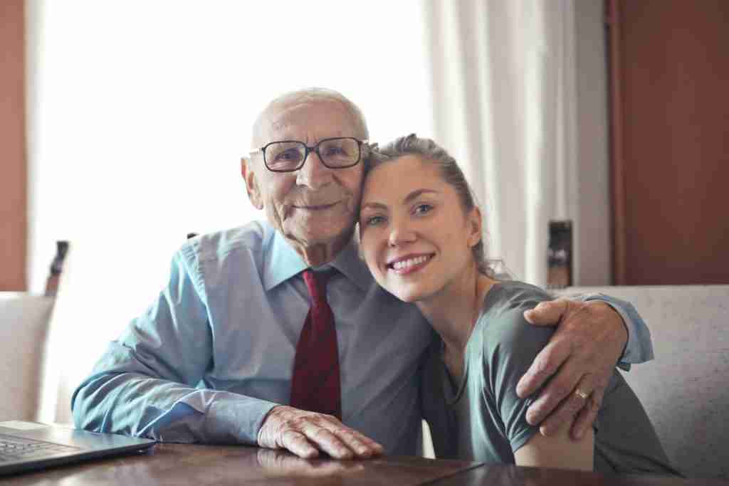 Positive senior man in formal wear and eyeglasses hugging with young lady while sitting at table