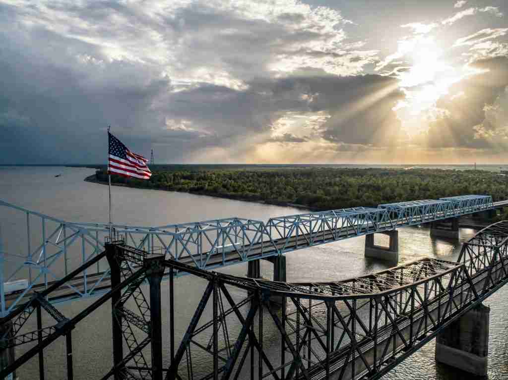 A scenic bridge under a cloudy sky during the day, featuring a flag with interesting facts about the Mississippi River.